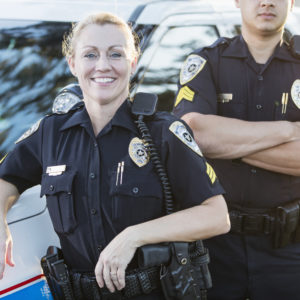 A policewoman standing beside her squad car, leaning against it, looking at the camera. She is a mature woman in her 40s. Her partner, an Hispanic man in his 30s, is standing behind her, cropped, with his arms crossed.